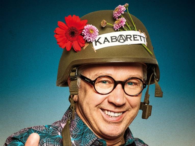 Lachender Mann mit Brille trägt einen Stahlhelm mit Blumen und der Aufschrift Kabarett.